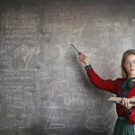 Serious female teacher wearing old fashioned dress and eyeglasses standing with book while pointing at chalkboard with schemes and looking at camera